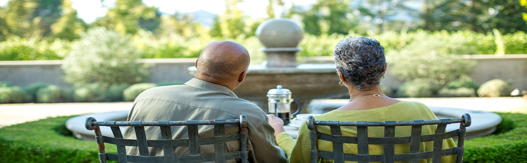 A view of the back of an older man and woman sitting on patio chairs outside in a garden.