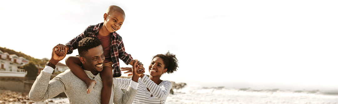 man smiling and carrying child on shoulders with woman looking at them and walking on the beach