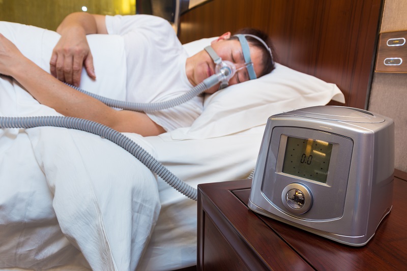 CPAP machine sitting on table next to a bed with sleeping man wearing headgear connected to air tube