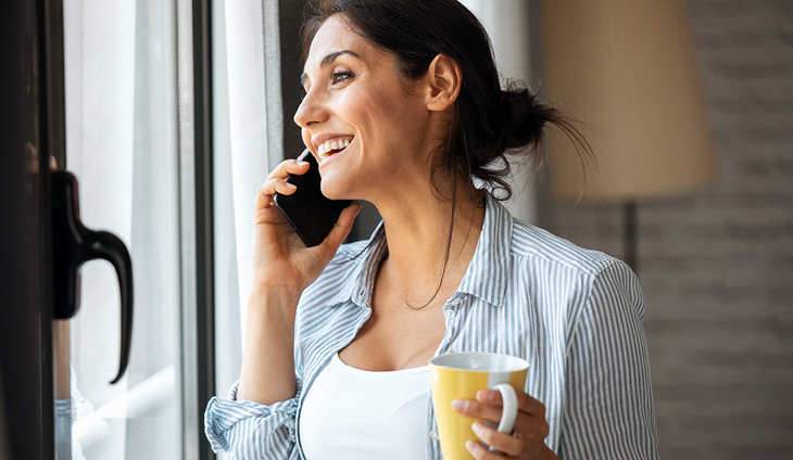 A woman looking out the window while holding a mug and talking on the phone