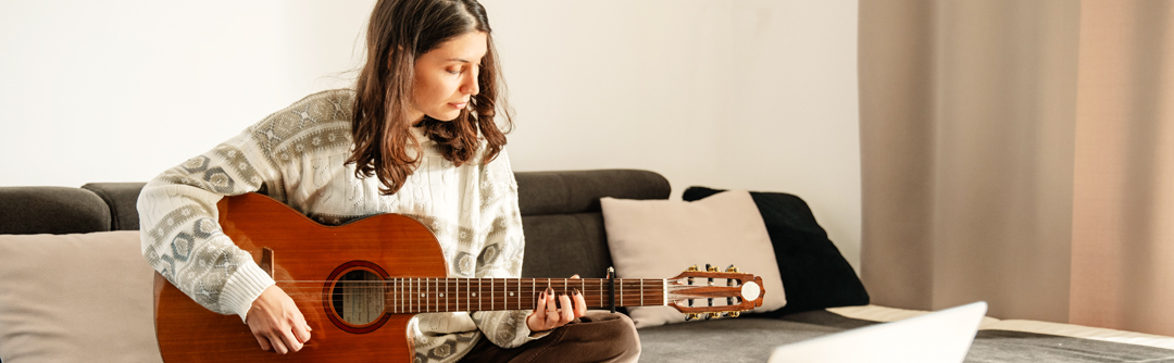A young woman sits on a couch playing the guitar, with her computer in front of her. 