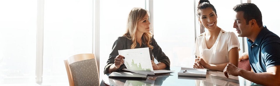 A smiling man and woman sit at a table with another woman, who is holding a document.