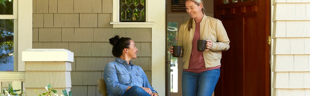 A young woman sits outside while another young woman brings over two coffee mugs.